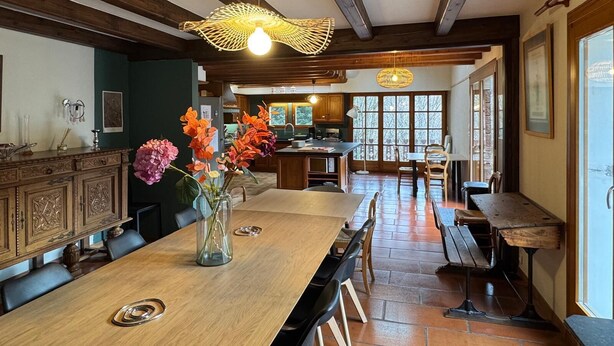 Een ruime eethoek met een lange houten tafel, stoelen en een vaas met bloemen. Achtergrond is een keuken met houten kastjes en een raam.