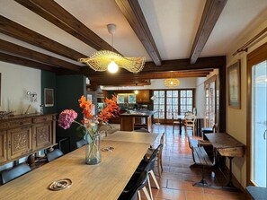 A spacious dining area with a long wooden table, chairs, and a vase of flowers. There is a kitchen in the background with wooden cabinets and a window.