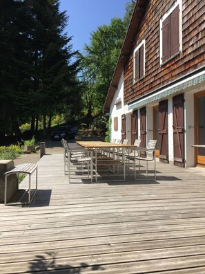 A wooden deck with a table and chairs, a car parked nearby, and a building with wooden shutters and a white facade.