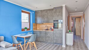 Cocina compacta con gabinetes grises, encimera de madera y horno empotrado. Cuenta con una zona de comedor con mesa y sillas, y un refrigerador pequeño.