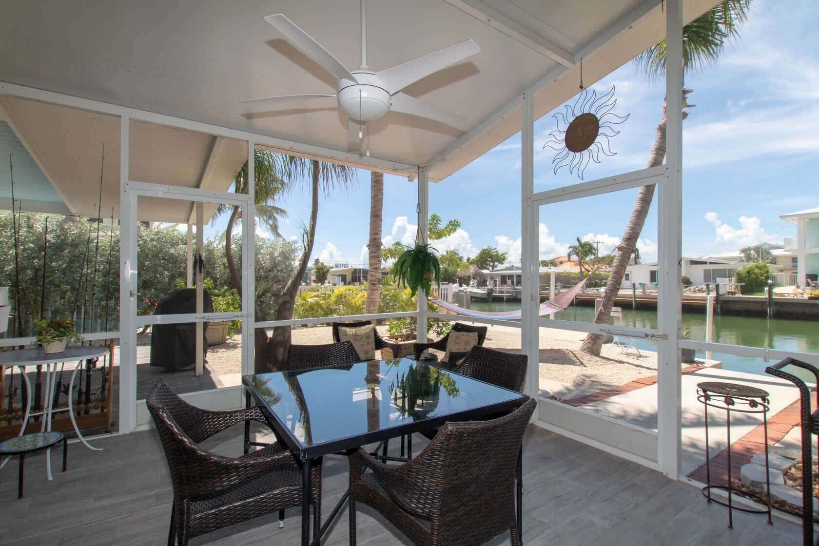 An outdoor dining area with a glass table and wicker chairs, overlooking a body of water and palm trees.
