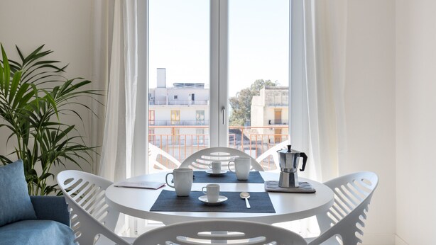 Ein Zimmer mit einem weißen Tisch für zwei Personen, einer Kaffeemaschine und Blick auf Gebäude durch große Fenster.
