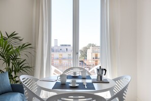 A room with a white table set for two, a coffee maker, and a view of buildings through large windows.