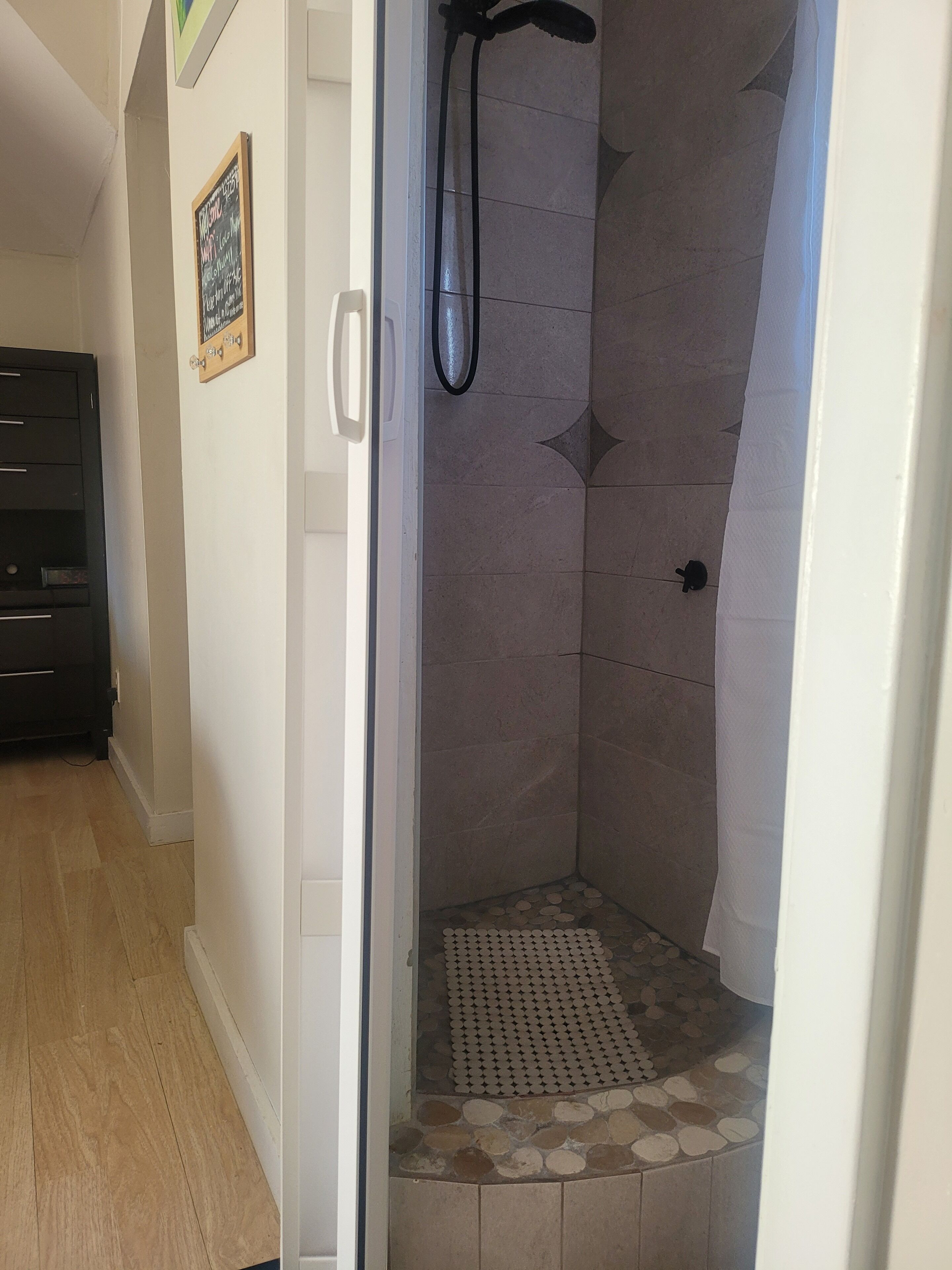 A shower with a rain shower head, a tiled floor with a pebble pattern, and a door leading to a hallway.