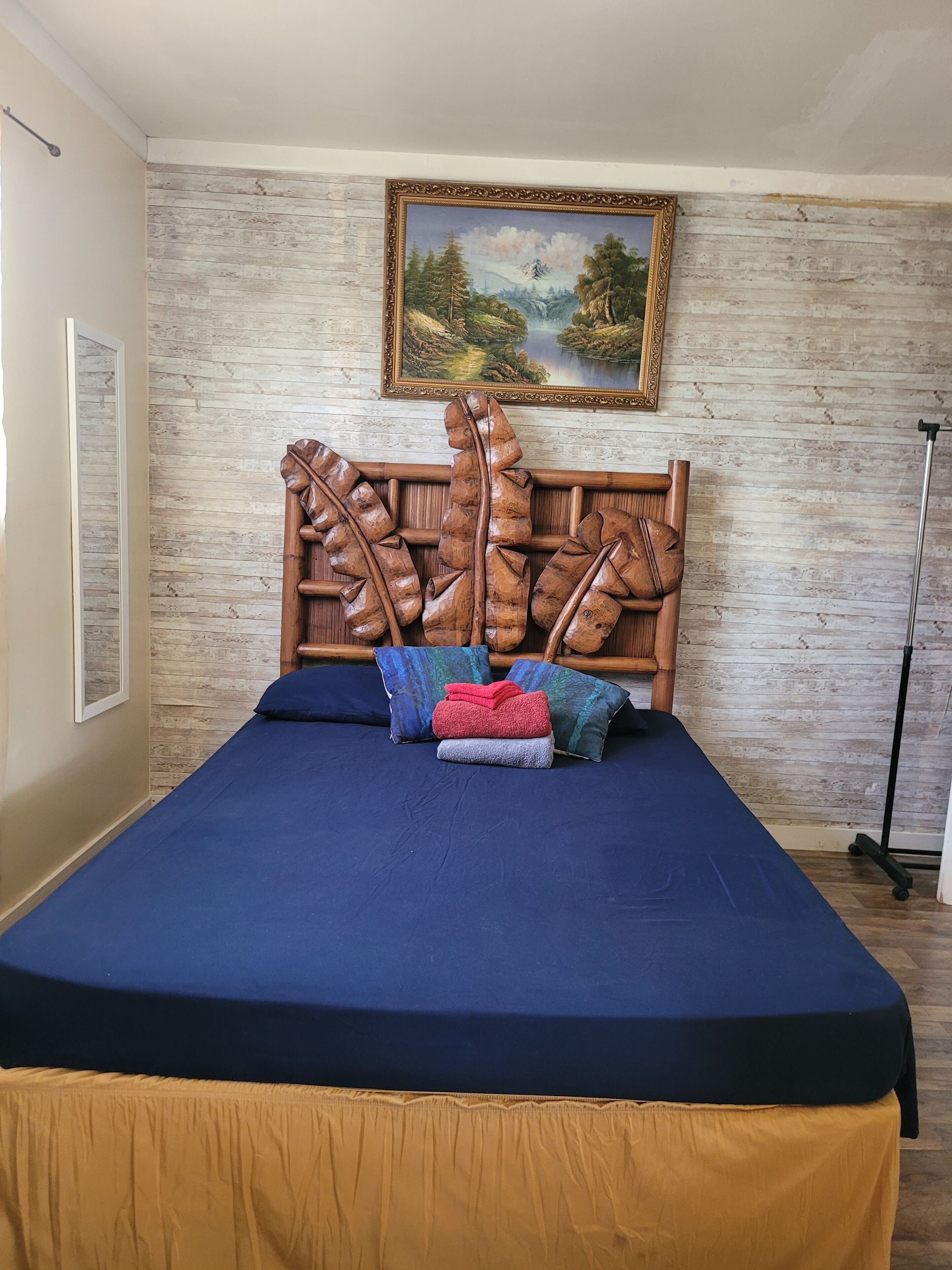 A bed with a blue mattress and a wooden headboard with a leaf design. There are pillows in red, blue, and gray. A painting of a landscape hangs above the bed.