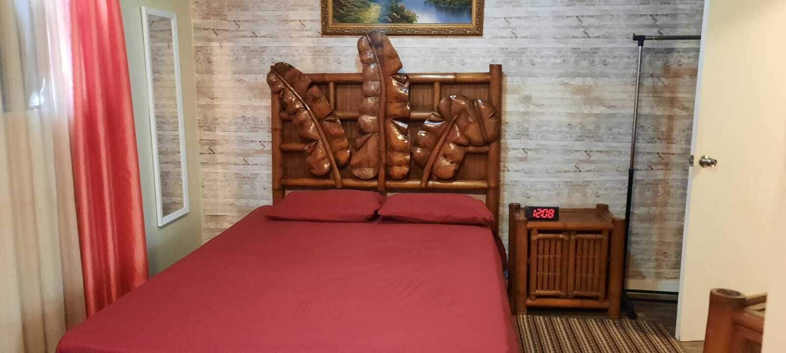 Una habitación con cabecera de madera, cama con ropa de cama roja, mesita de noche de madera y un reloj marcando las 12:08.