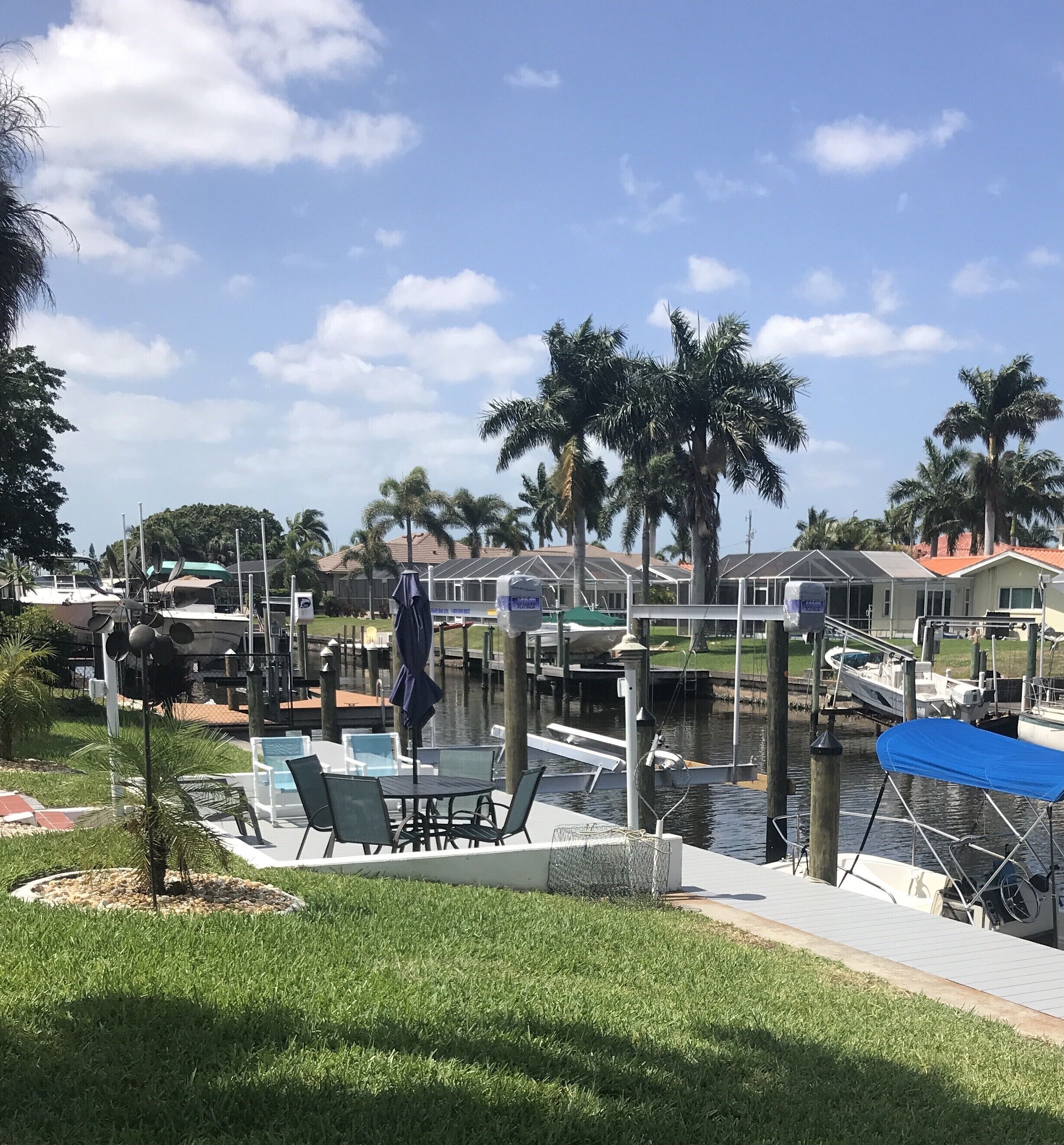Un port de plaisance avec des bateaux amarrés, une zone herbeuse avec des tables et des chaises, et des palmiers.
