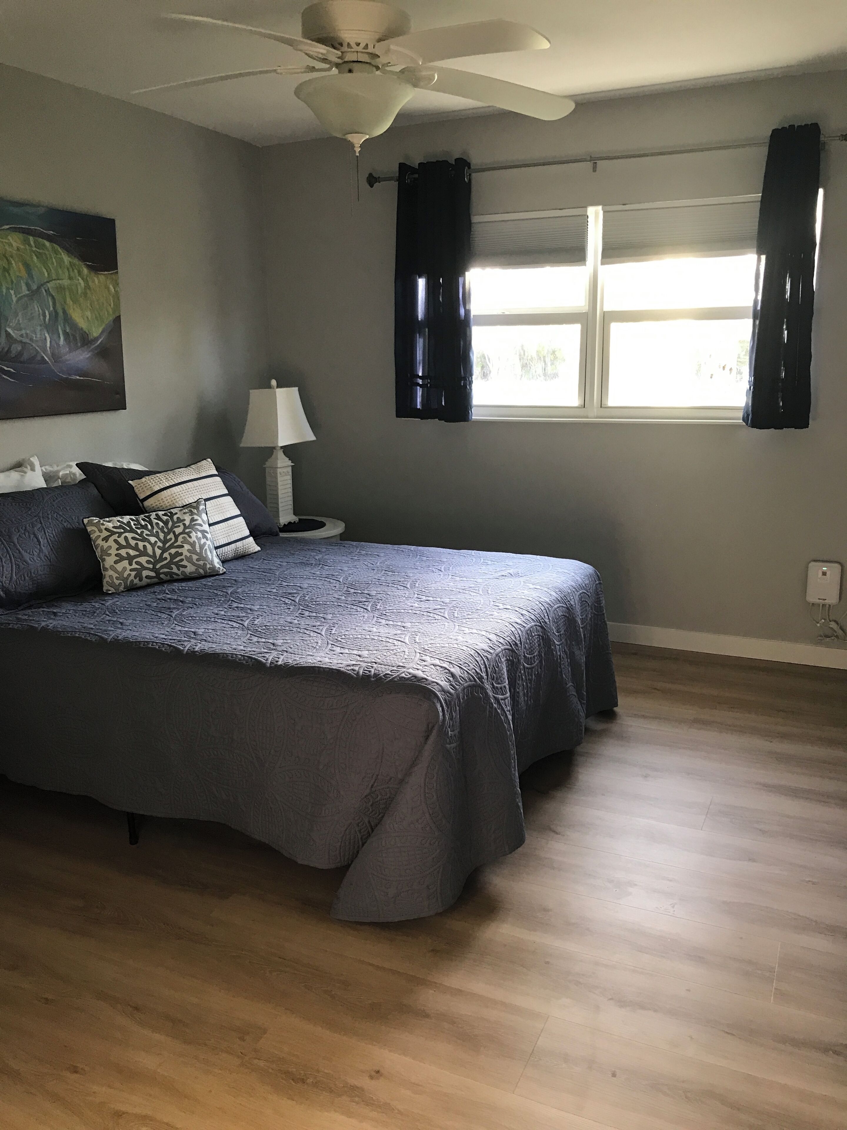 A bedroom with a bed, a ceiling fan, a window with curtains, and a painting on the wall.