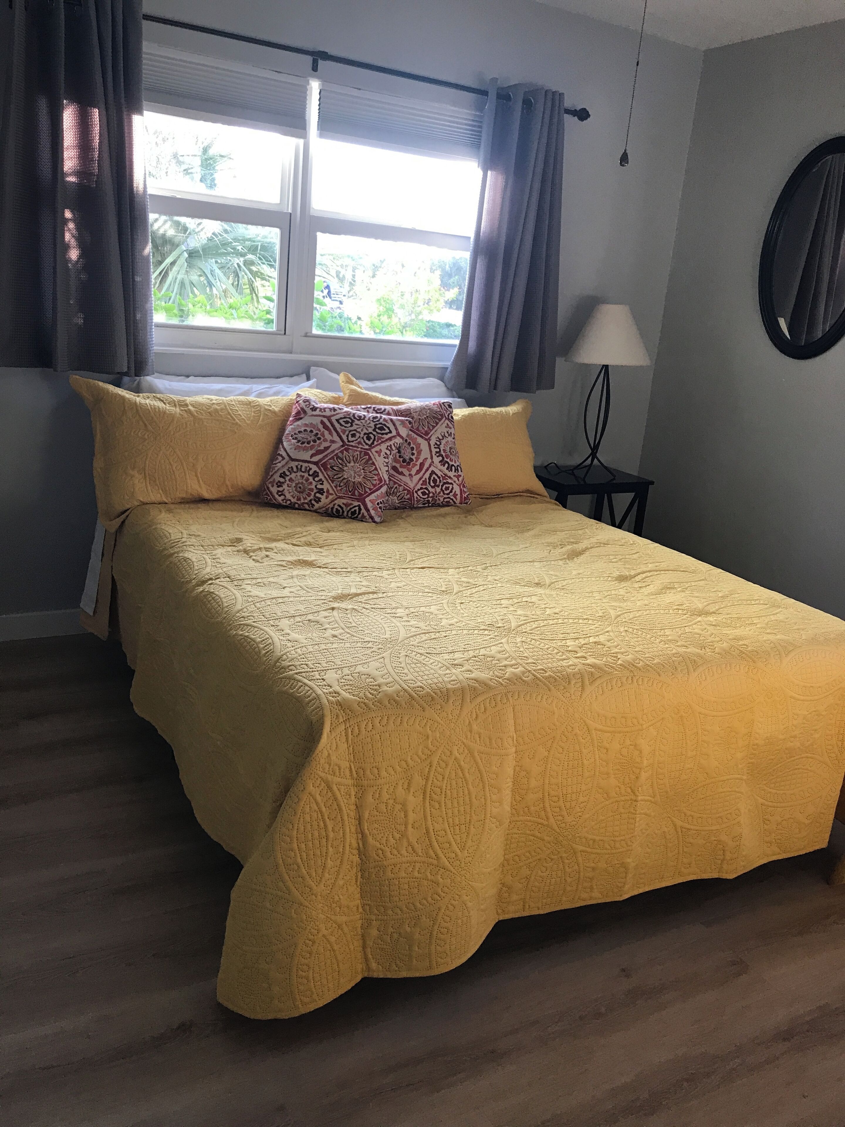 A neatly made bed with a yellow bedspread and patterned pillows, a small bedside table with a lamp, and a window with curtains.