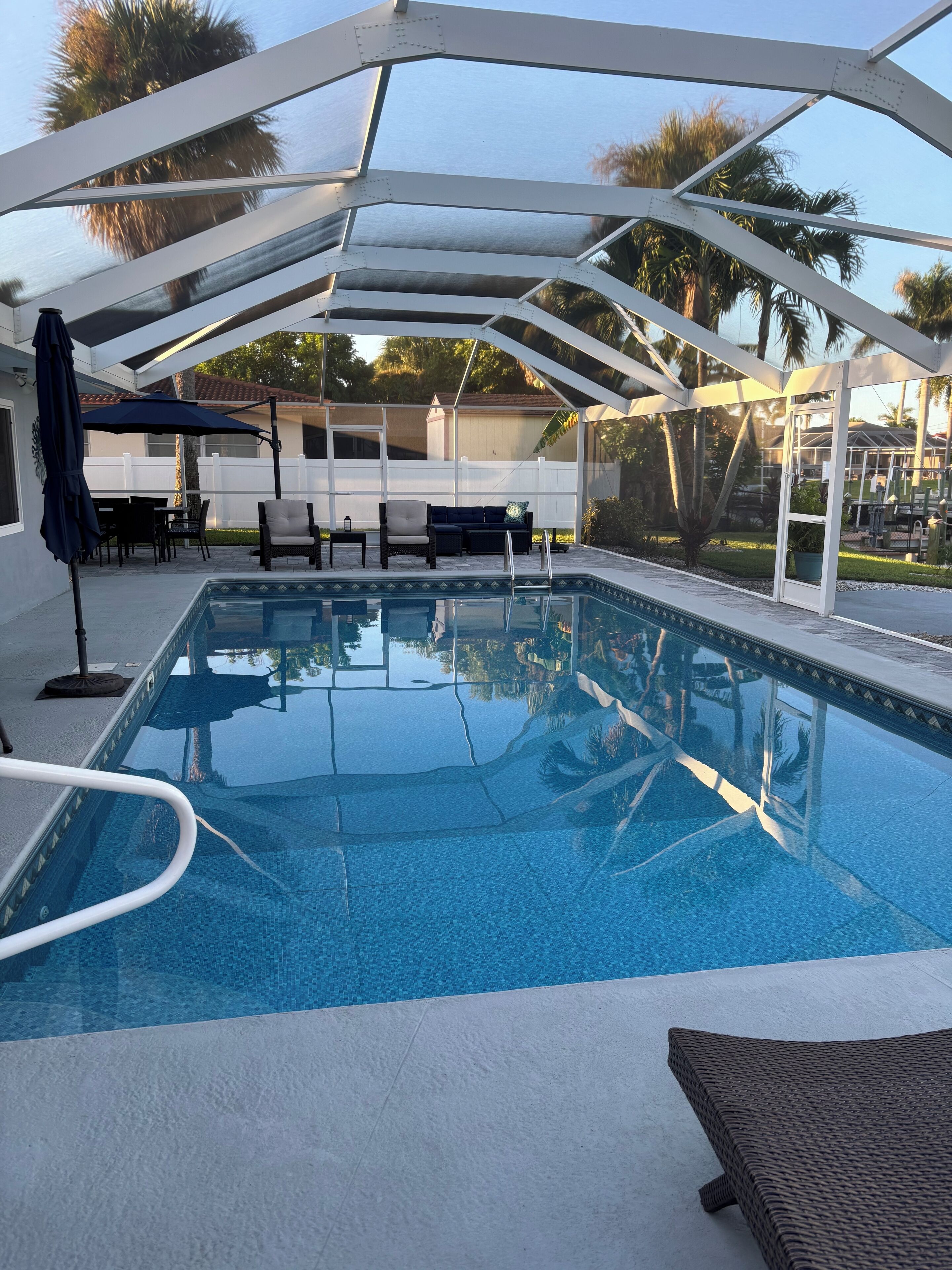 A covered swimming pool with lounge chairs and umbrellas, surrounded by a white fence and palm trees.