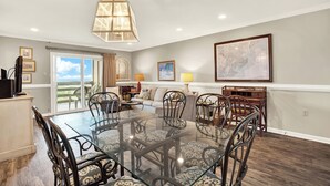 A spacious dining area with a glass table and metal chairs, a large chandelier, and a view through a sliding glass door to the outdoors. - Experience Lighthouse Point 35C– Your North End Tybee Retreat (Tybee Island)