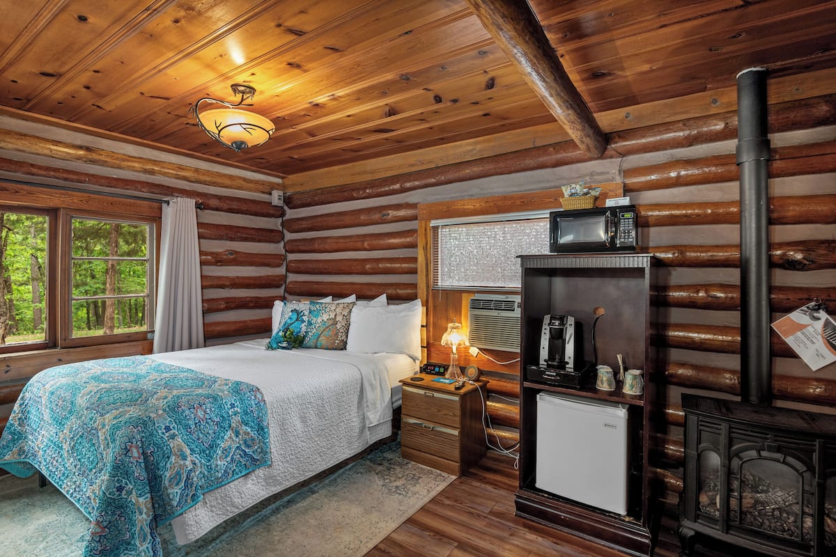 A log cabin bedroom with a bed, bedside table, lamp, and a wood-burning stove.