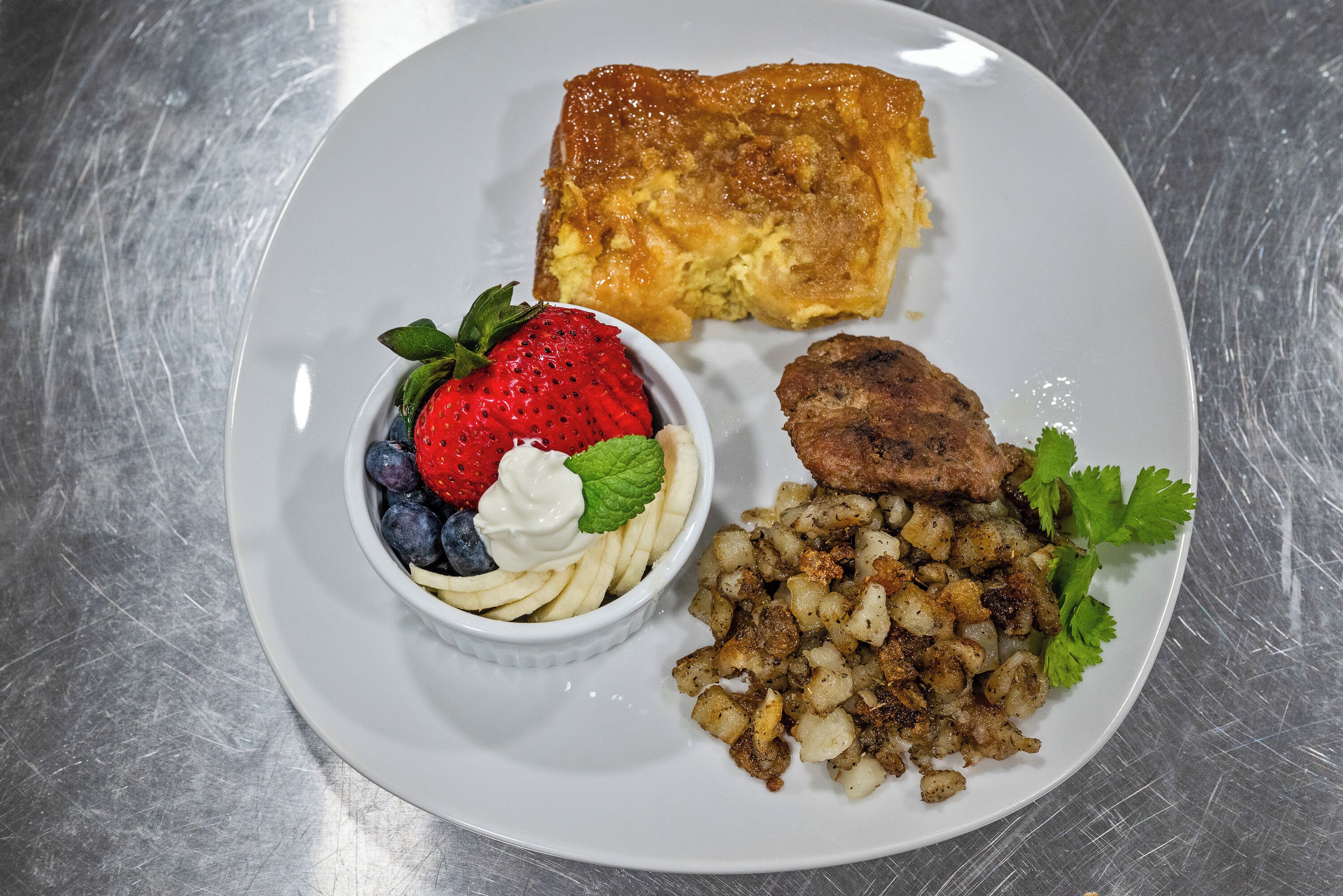 Une assiette contenant une portion de plat cuit au four, doré et croustillant, un petit bol de fruits et de crème fouettée, ainsi qu’une portion de pommes de terre assaisonnées.
