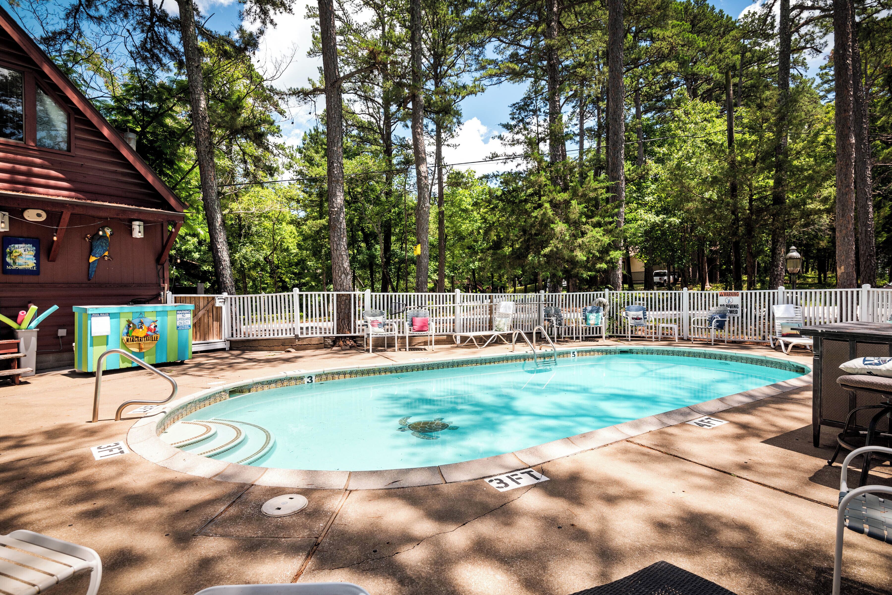 A swimming pool with a wooden deck, surrounded by a white fence, and trees in the background.