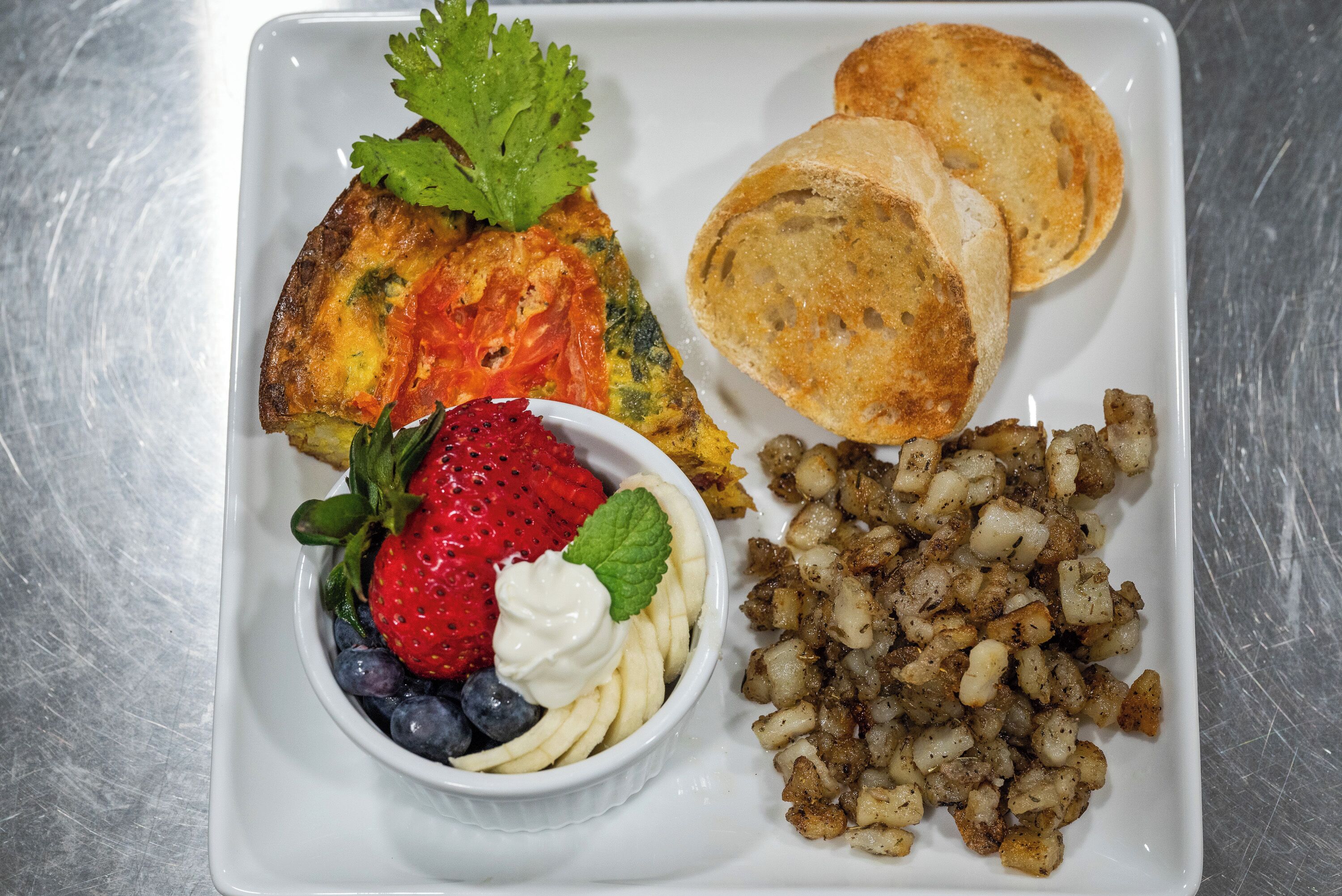 Une assiette de petit-déjeuner comprenant des toasts, des œufs brouillés, des pommes de terre rissolées et un saladier de fruits frais, avec des fraises et des myrtilles.