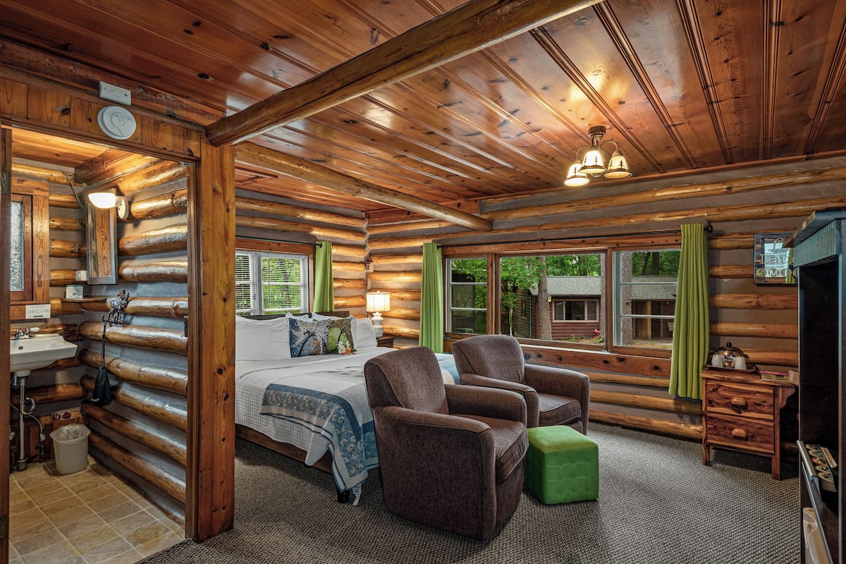 A log cabin interior with a bed, two armchairs, a green ottoman, a wooden dresser, and a window with curtains.