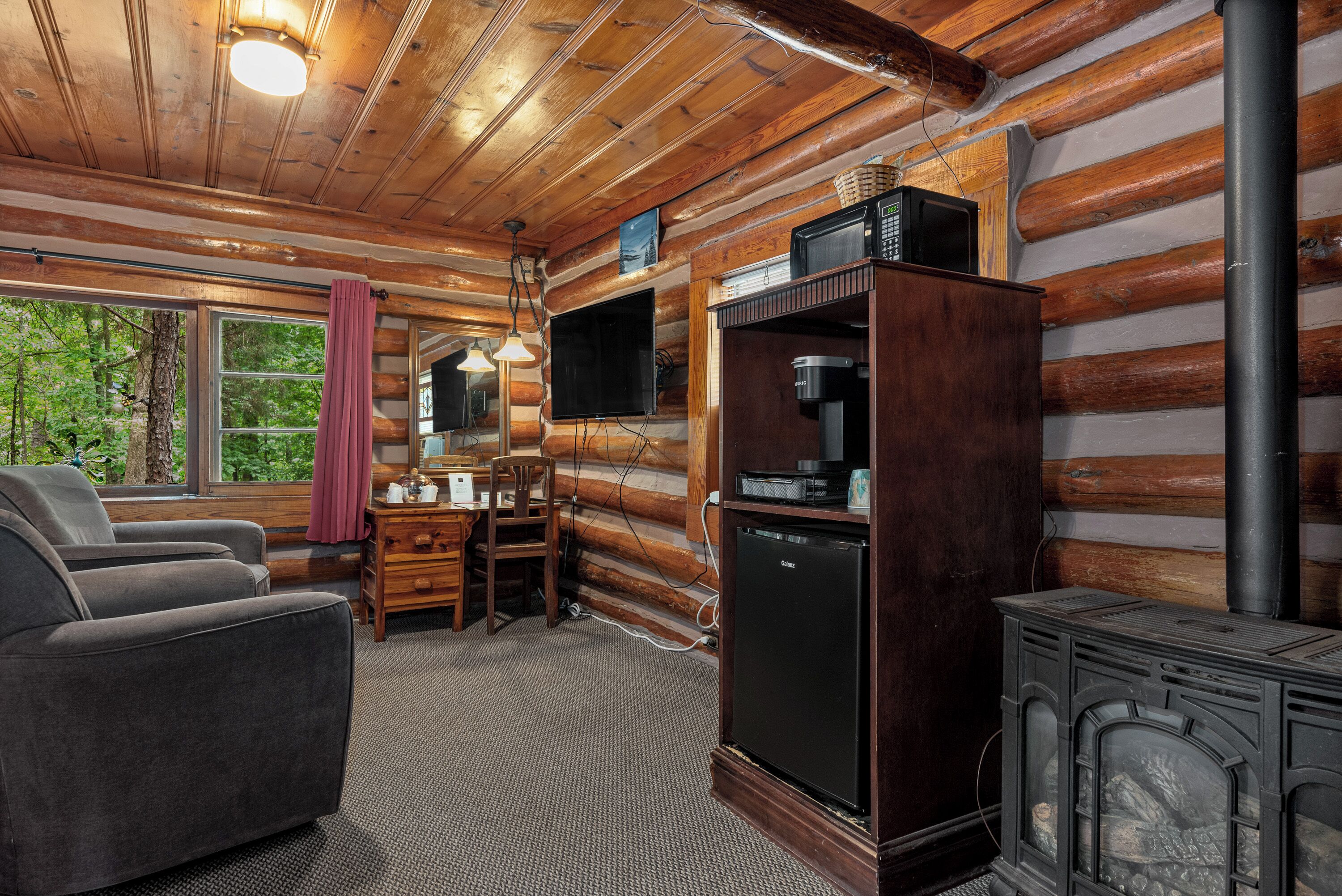 Interior de una cabaña de troncos con sofá, escritorio de madera, televisor y estufa de leña.