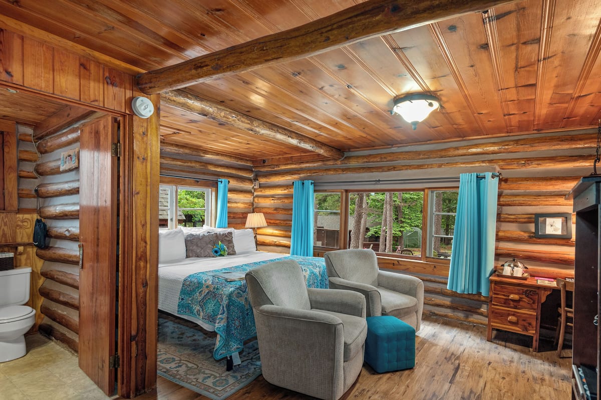 A log cabin bedroom with a bed, two armchairs, a wooden dresser, and a window with curtains.