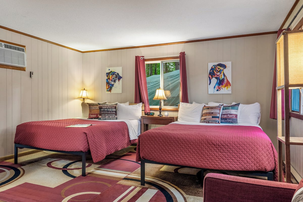 A hotel room with two beds, a window with curtains, a lamp, and artwork on the walls.