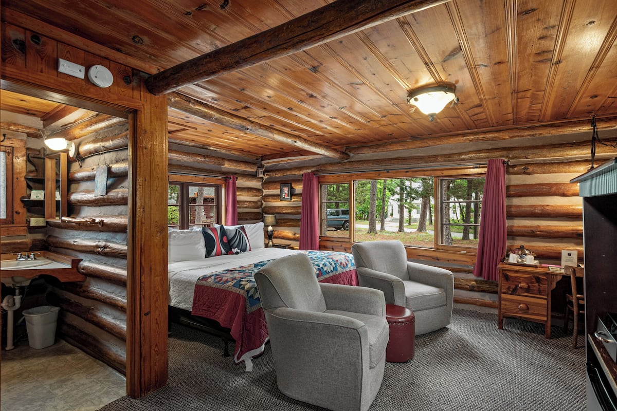A log cabin room with a bed, two armchairs, a desk, and a window with curtains.