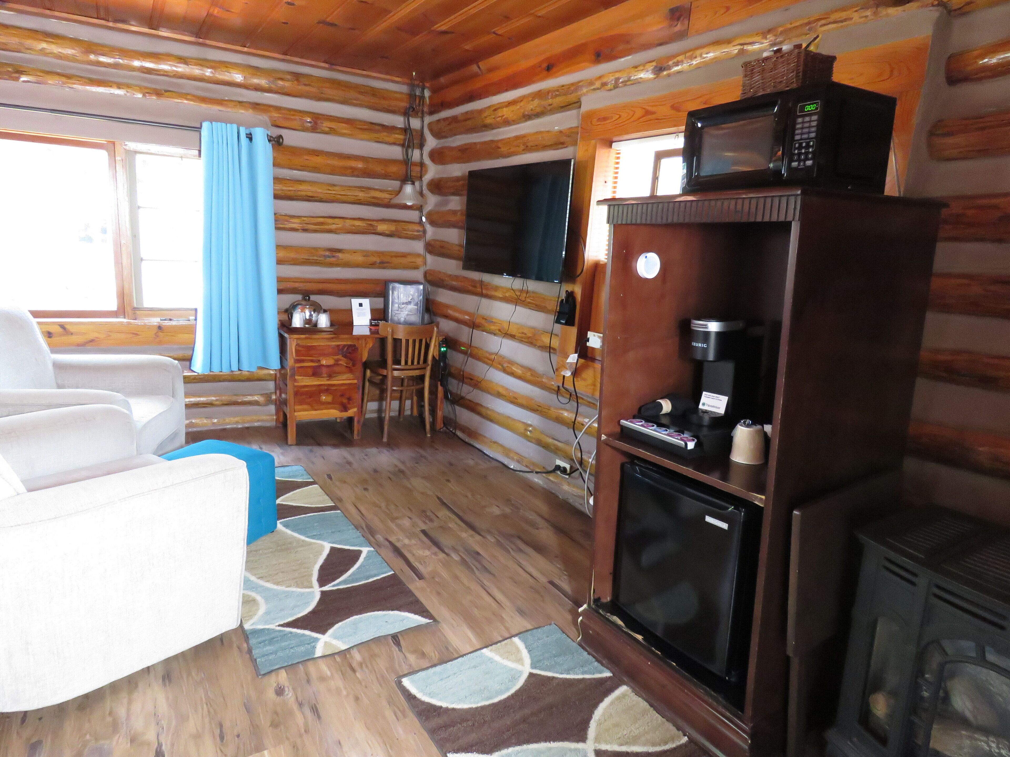 Interior de una cabaña de troncos con un escritorio de madera, un sillón blanco, un televisor, una cafetera y un estufa de leña.