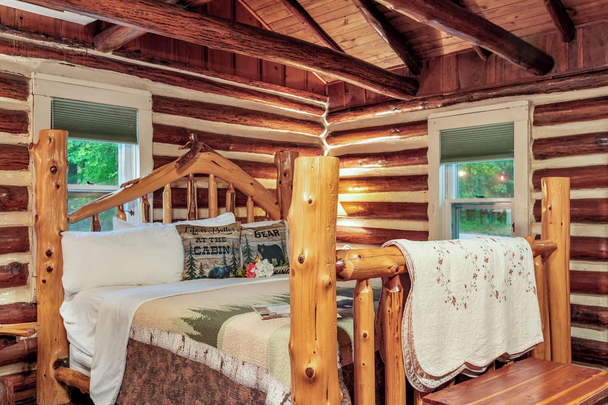 A log cabin room with a wooden bed, a window with a view of trees, and a quilt with a bear design.