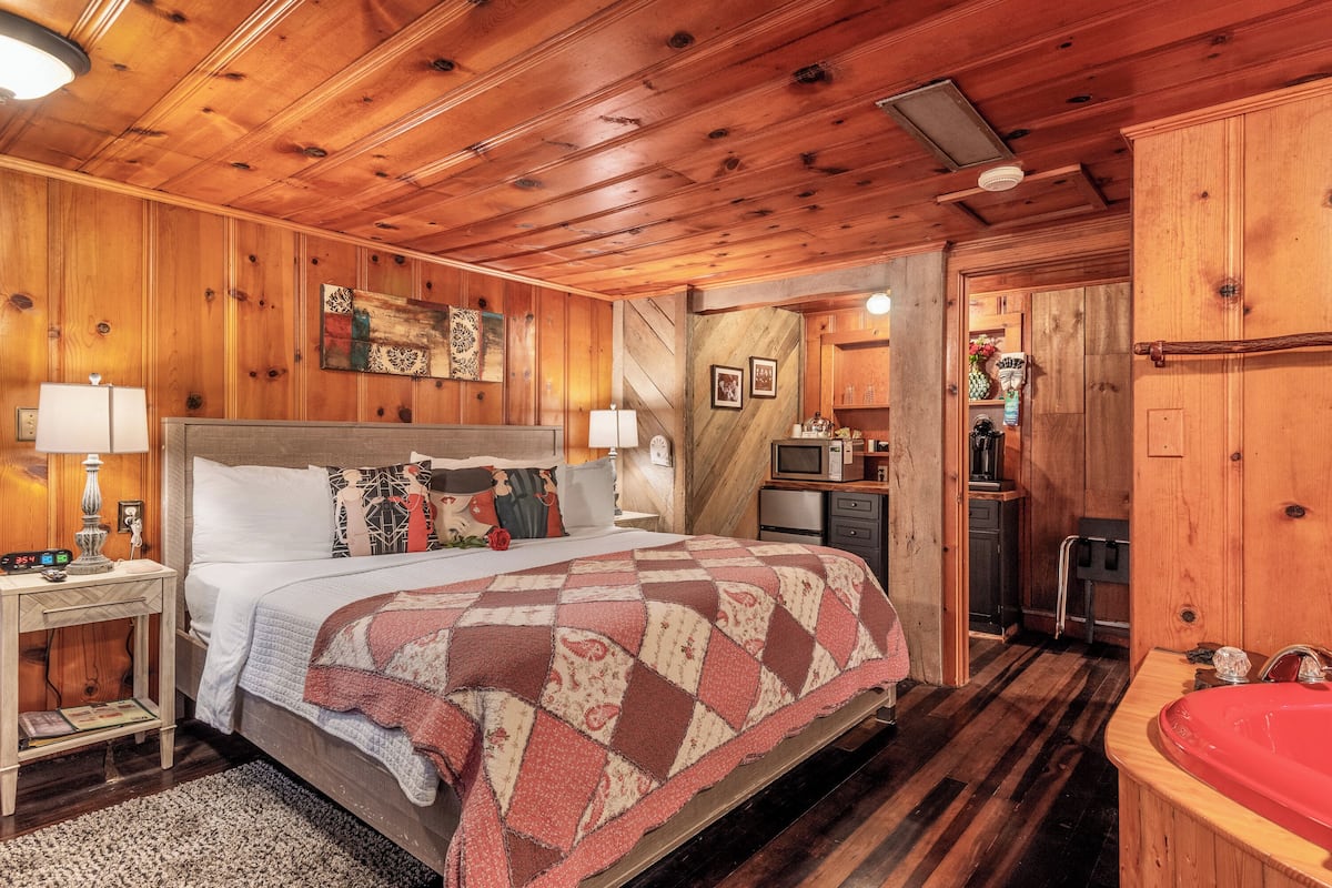 A wooden cabin bedroom with a bed, bedside tables, a red bathtub, and a small kitchenette.