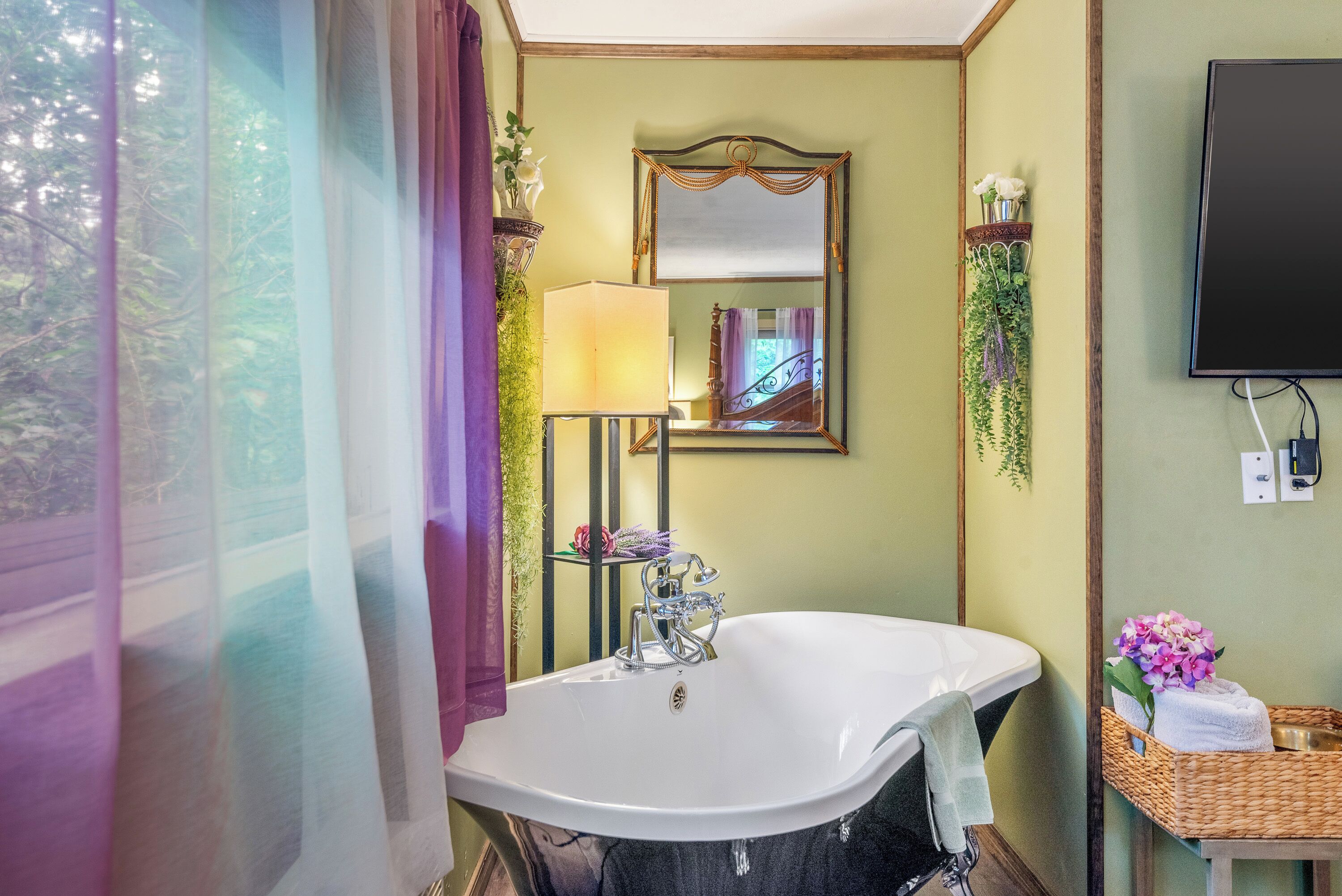 Une salle de bain avec une baignoire sur pieds, un miroir, un téléviseur fixé au mur et un panier en osier contenant des serviettes.