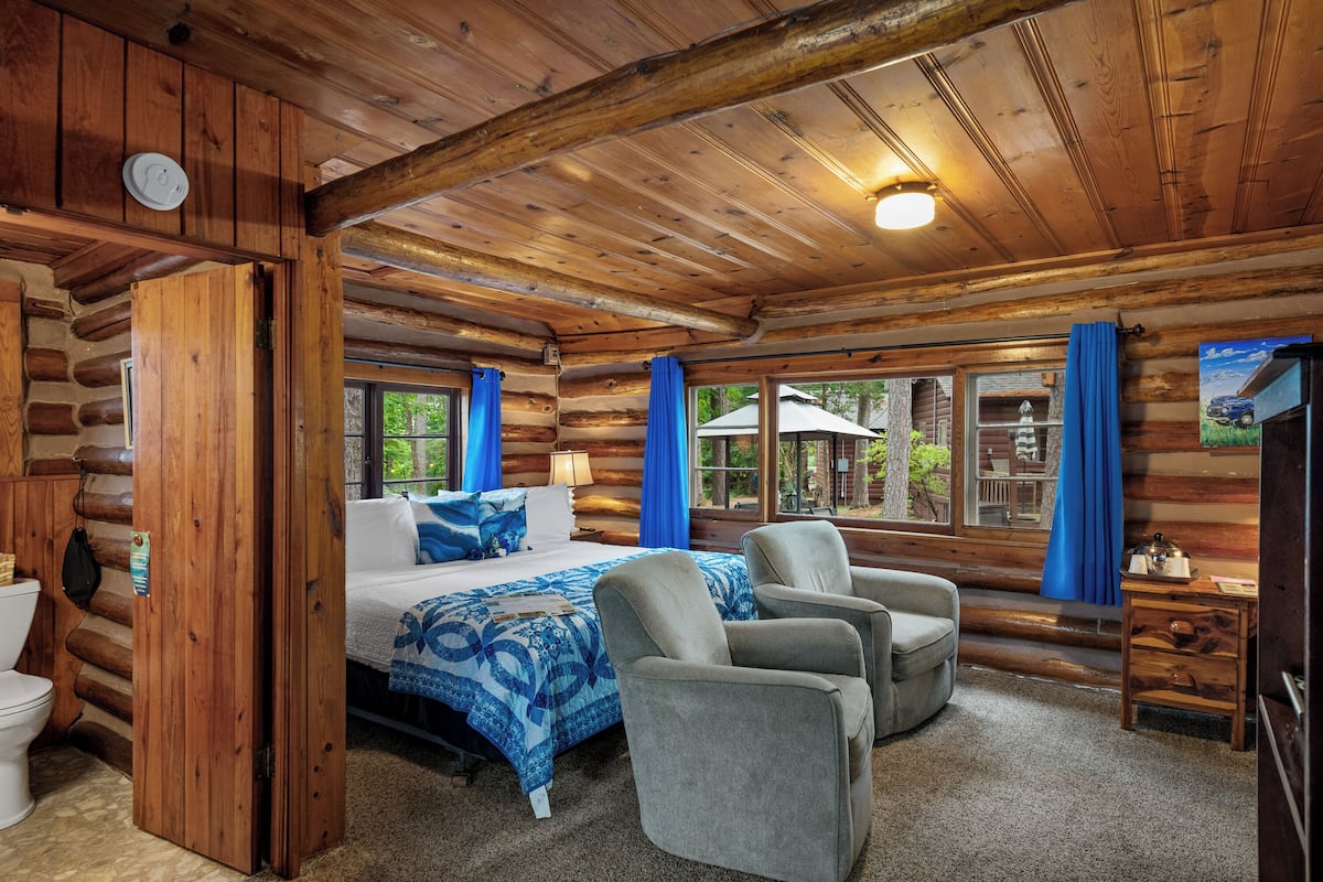 A log cabin bedroom with a bed, two armchairs, a wooden nightstand, and a window with curtains.