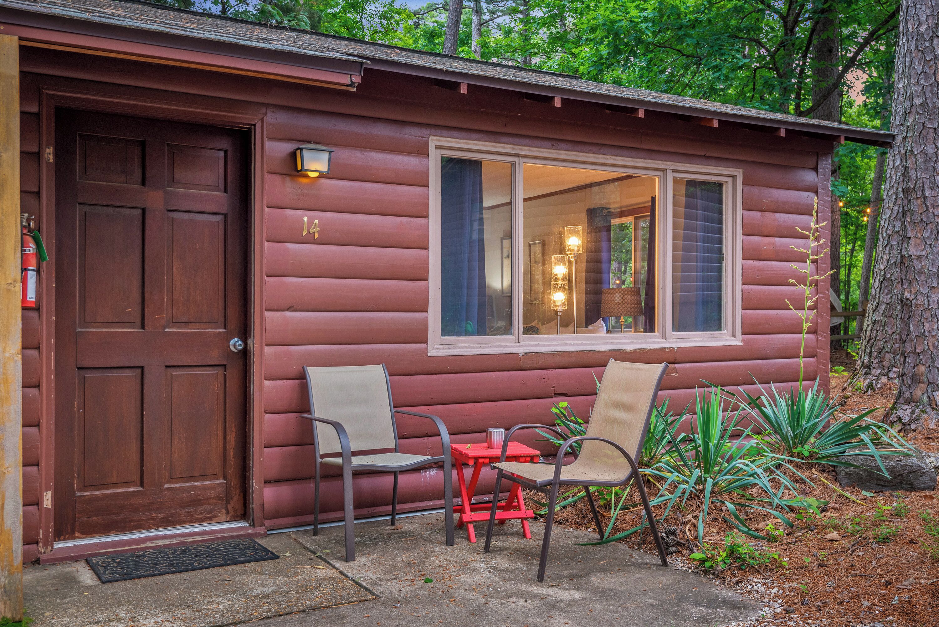 A log cabin with a wooden door, a window with a view of the interior, and two chairs outside.