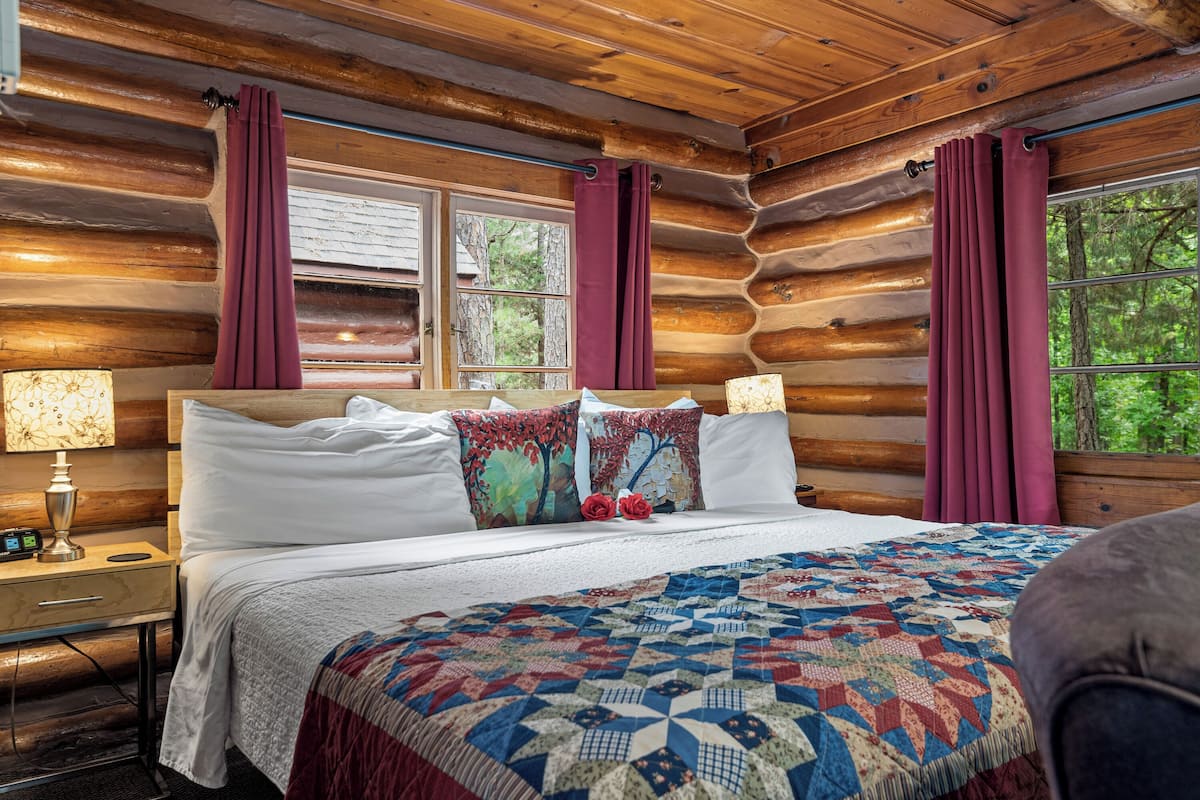 A log cabin bedroom with a large bed, two pillows, a quilt, and a nightstand with a lamp.
