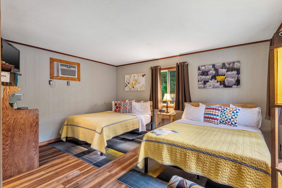 A hotel room with two beds, a wooden desk, a window with curtains, and artwork on the walls.