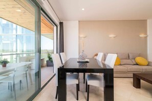 A modern living room with a glass partition, a dining table with chairs, a sofa with cushions, and a decorative vase. - Marina Vilamoura - Aquamar 401- Modern Apartment (Quarteira)