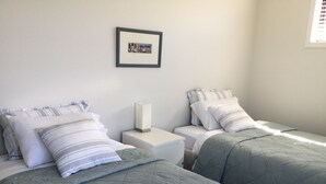 A bedroom with two single beds, each with a grey quilted duvet and striped pillows. There is a bedside table with a white lamp between the beds. A framed picture hangs on the wall above the beds. - Northlake Wanaka New Zealand. House to let in Ski Season. (Wanaka)