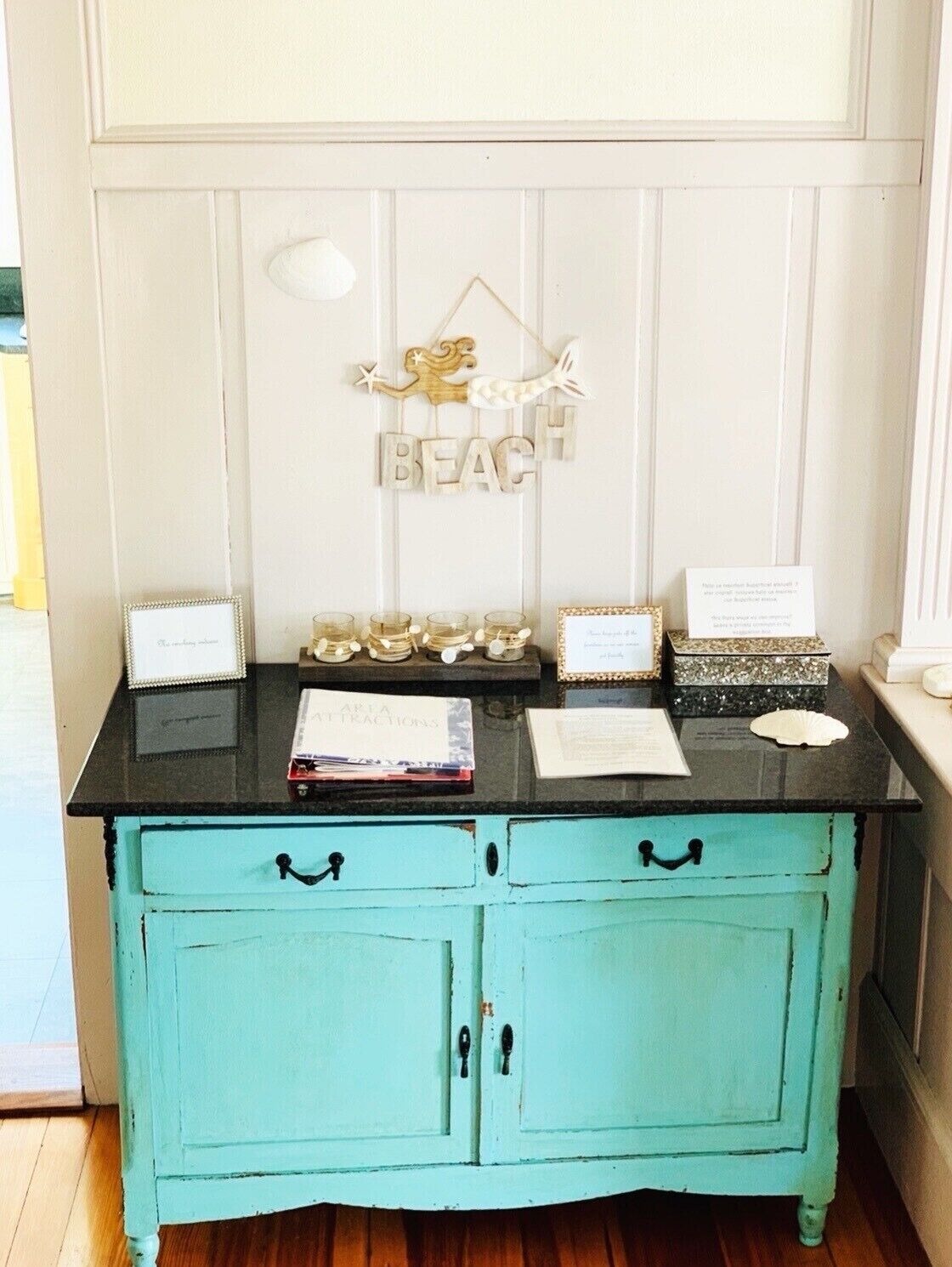 A teal-painted wooden cabinet with two drawers and a black countertop. Above the countertop, there are decorative items including a sign that reads "BEACH" and several small jars.