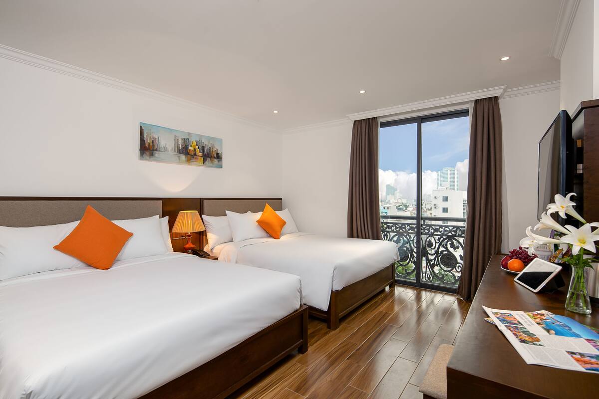 A hotel room with two beds, a balcony with a city view, a desk with a laptop, and a vase with flowers.