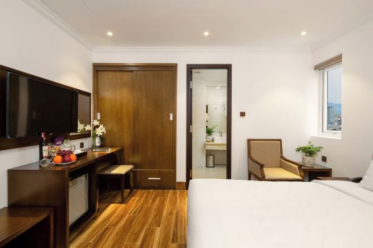 A hotel room with a bed, a desk with a flat-screen TV, a chair, and a window.