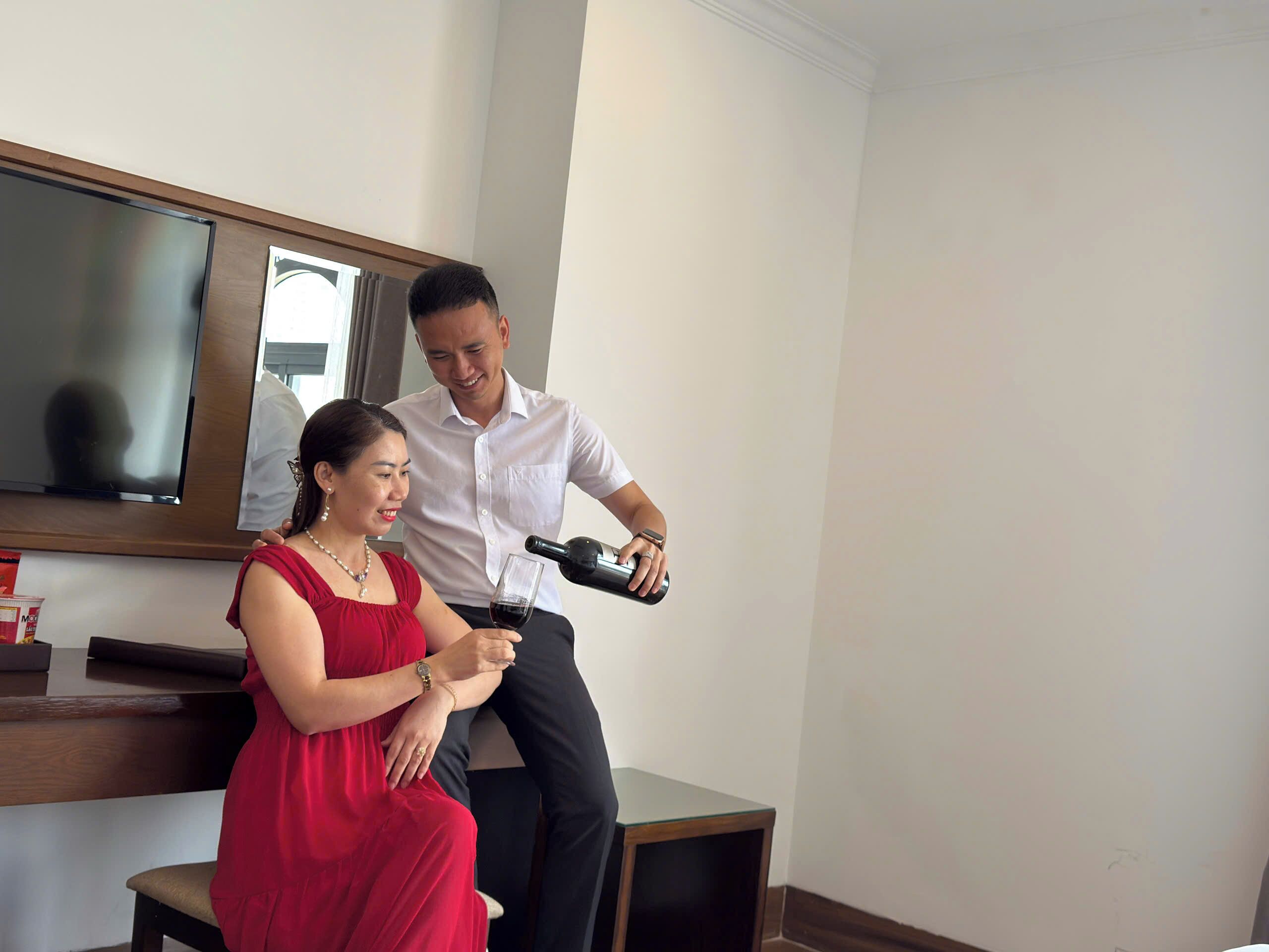 A man pouring a drink into a glass, a woman seated on a chair, a television mounted on the wall, and a wooden table with a remote control.