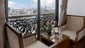 Un balcón con vistas a un paisaje urbano, dos sillones tapizados, una mesa de vidrio con una botella y un vaso, y un florero con flores.