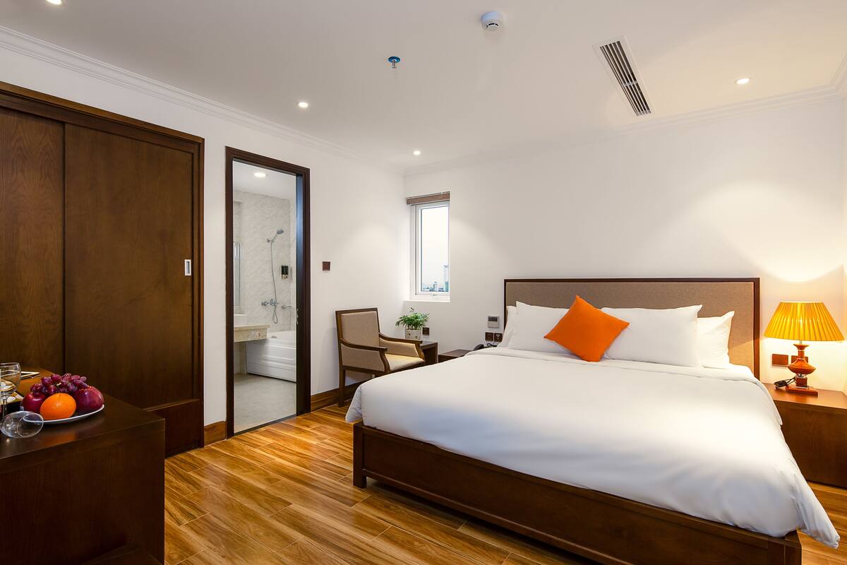 A hotel room with a wooden bed, a bedside table with a lamp, a desk with fruit and glasses, and a view of the bathroom.