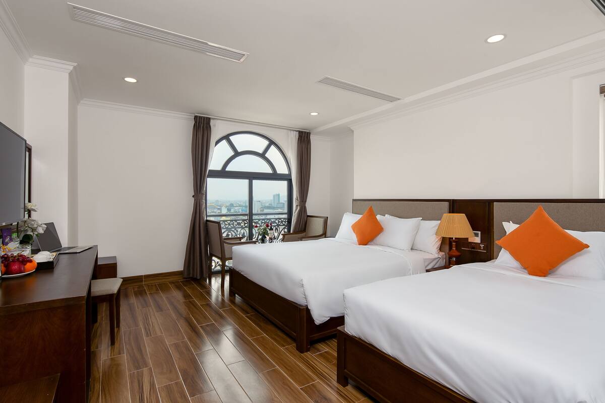 A hotel room with two beds, a desk, a chair, and a view of a cityscape through a large window.