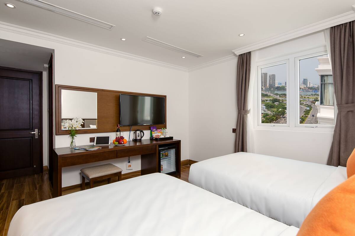 A hotel room with two beds, a desk with a computer, a small refrigerator, a TV, and a window with a city view.