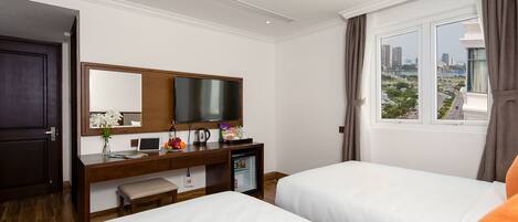 A hotel room with two beds, a desk with a computer, a small refrigerator, a TV, and a window with a city view.