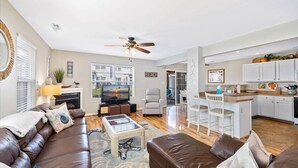 A spacious living room with a brown leather sofa, a coffee table, and a television. There is a kitchen area with white cabinets and a dining table.