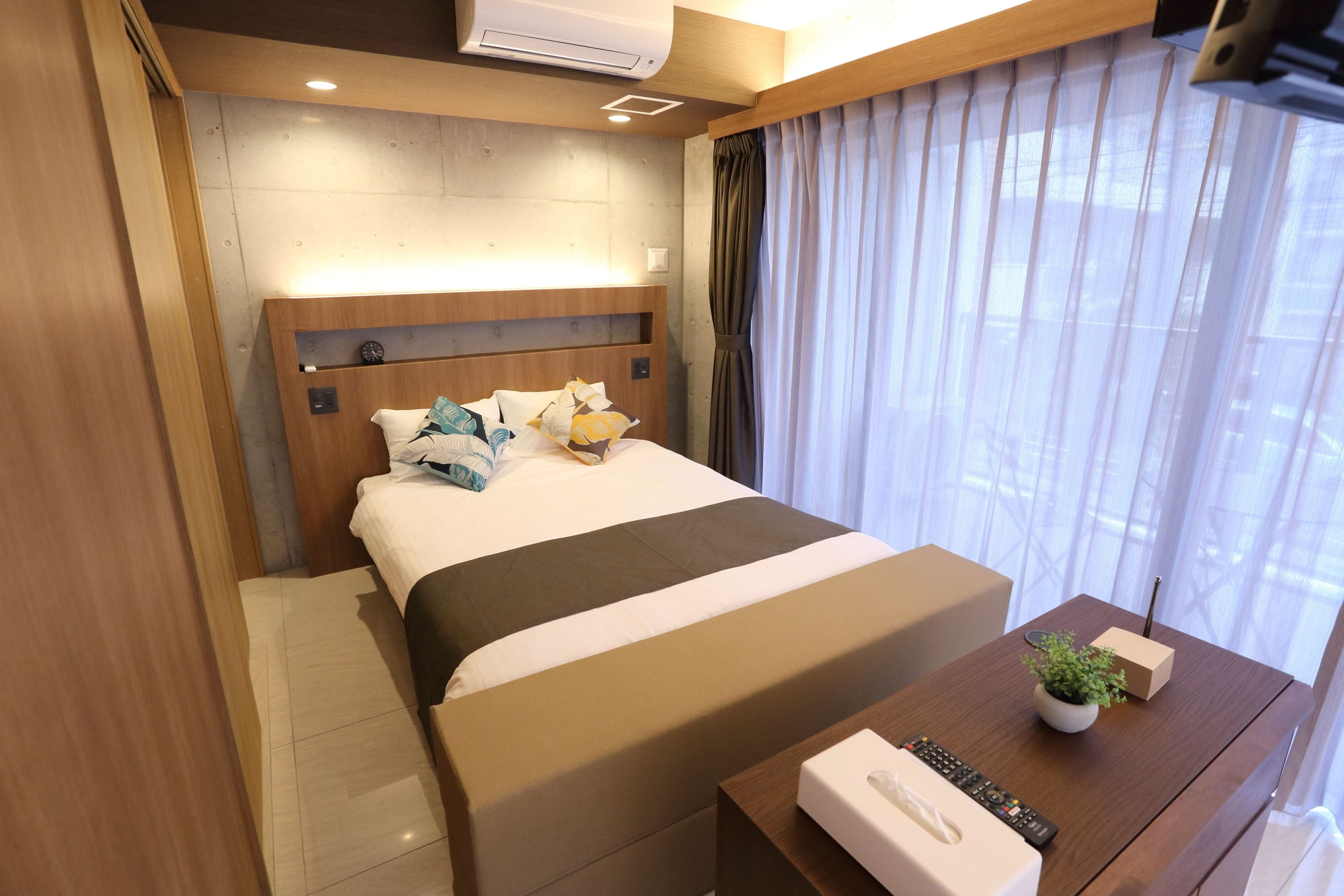 Una habitación de hotel moderna con una cama grande, cabecera de madera, mesita de noche con un control remoto, una planta pequeña y una ventana con cortinas translúcidas.