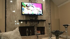 A living room with a flat-screen TV mounted on a wall, a fireplace, and a dining area with a table and chairs. - “NolaNeedeD Estate🏡” For Fun Vacations (New Orleans)