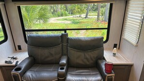 Interior acolhedor de uma RV com dois assentos reclináveis, uma pequena mesa com abajur e uma janela com vista para um jardim.