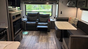Interior compacto de um veículo recreativo, com cozinha compacta, área de estar e janela com vista para a vegetação.