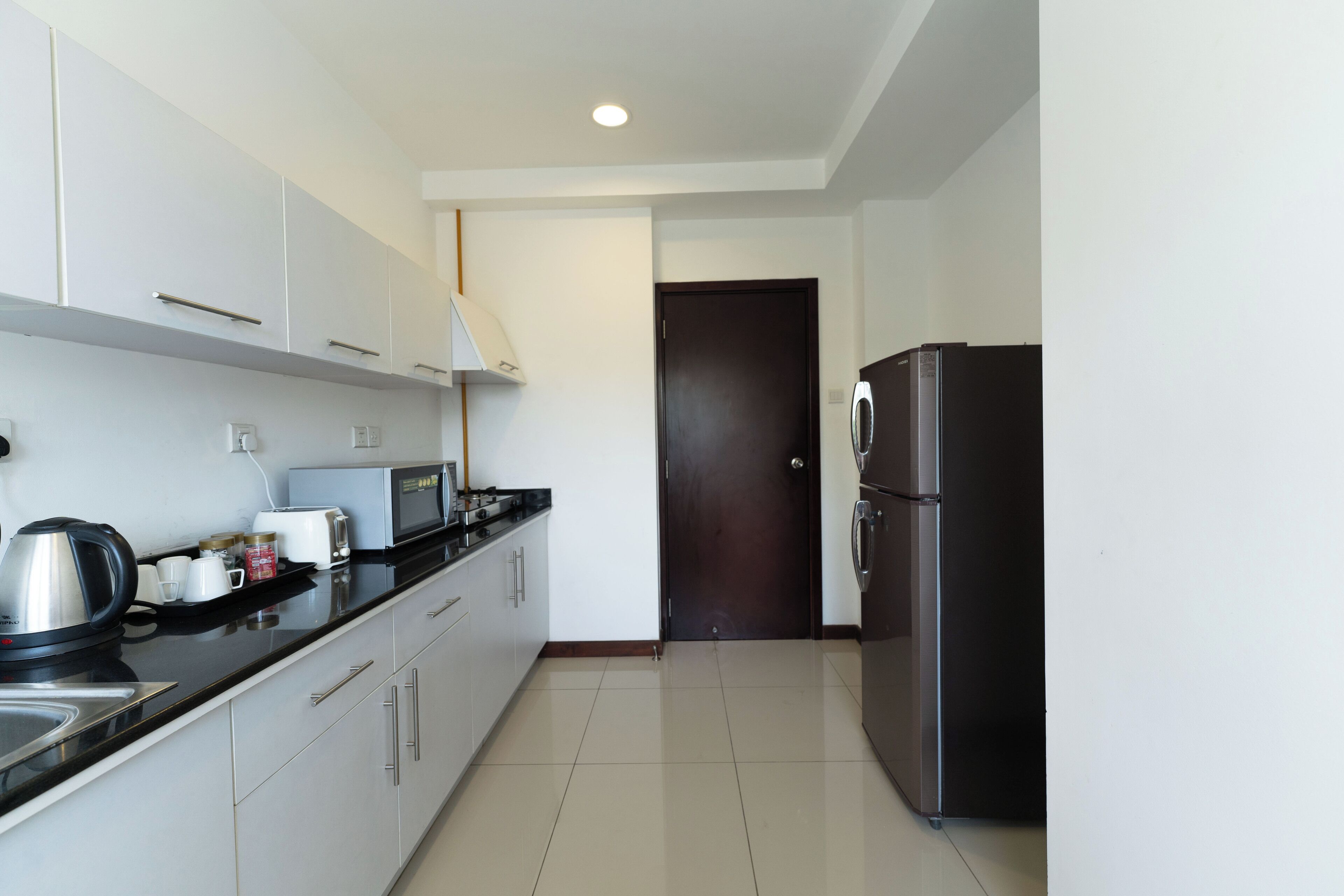 A modern kitchen with white cabinets, a black countertop, a microwave, a kettle, and a refrigerator.