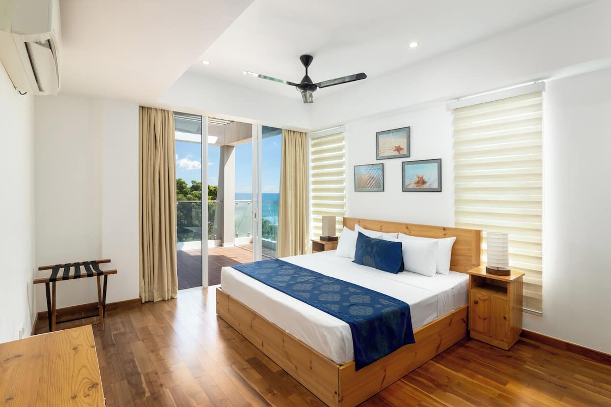 A bedroom with a large bed, wooden headboard, and a ceiling fan. There is a balcony with a view of the sky and trees.